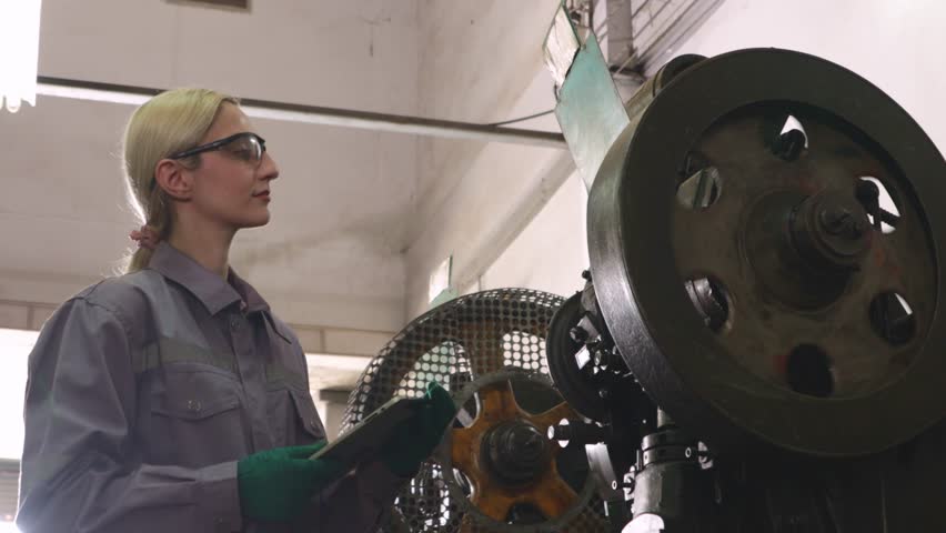 Female lathe operator wearing safety glasses chips and holding inspection tablet while carefully controlling metal stamping machine working with thin metal sheet in parts manufacturing plant.