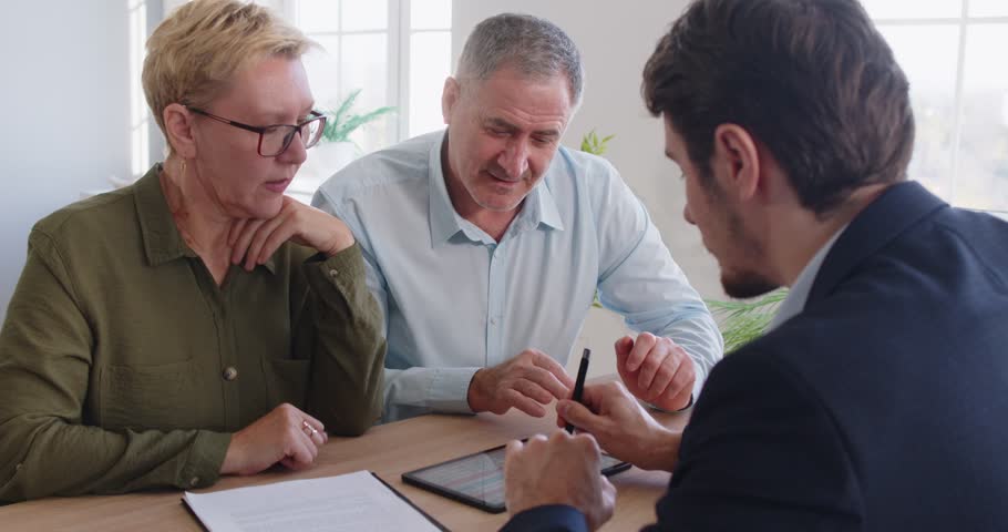 Male financial advisor meeting with senior family couple, sitting at desk and looking at tablet screen. Positive pensioners having consultation, discussing investment and life insurance decisions.