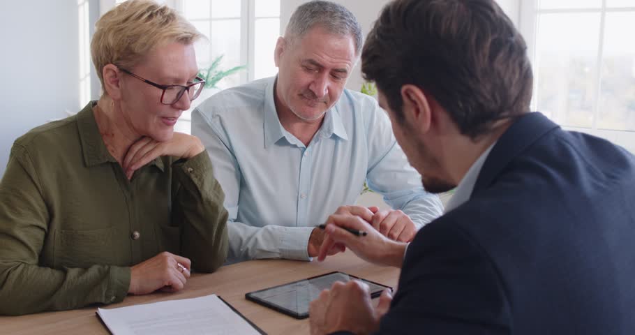 Male financial advisor meeting with senior family couple, sitting at desk and looking at tablet screen. Positive pensioners having consultation, discussing investment and life insurance decisions.
