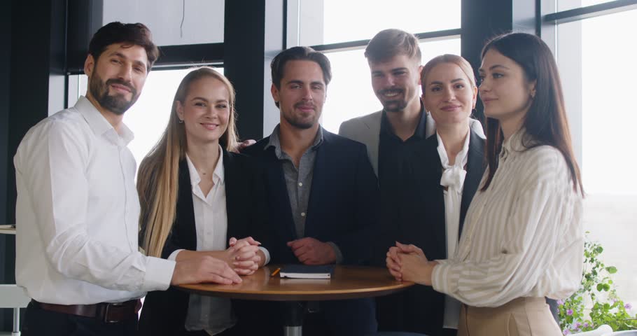 Happy cheerful business people men and women looking at camera hugging and smiling indoors standing at workplace. Company employees after meeting in office. Teamwork concept. 4k video.