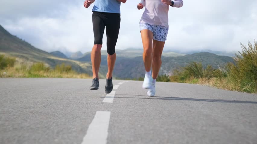 The muscular, powerful legs of a male and female athlete pound a deserted road at a fast pace. Scenic mountain views surround them, providing perfect conditions for an intense workout.
