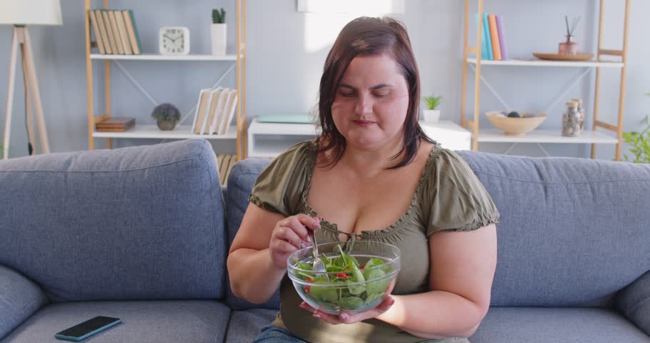 Upset overweight woman sitting on sofa at home and eating fresh vegetable salad. Unhappy plus size female looking thoughtful and showing dissatisfaction while trying to adopt healthy eating habits.