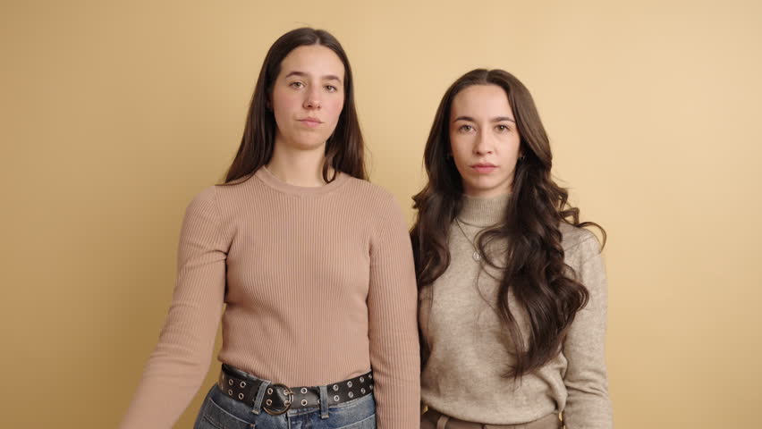 Real time young female friends with twisted mouths looking at camera while standing with stretched arms with thumbs down gesture and nodding heads sideways against beige background