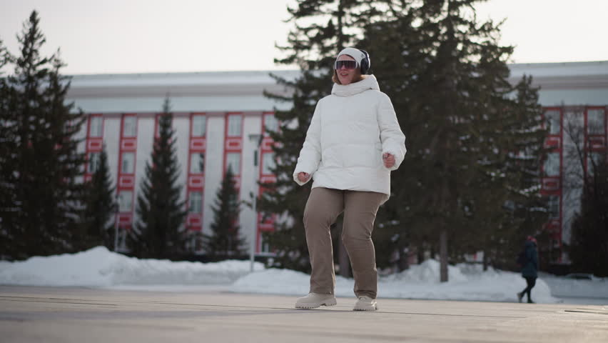 Closeup of young woman wearing headphones, beanie, goggles and puffer jacket dancing energetically on tiled street plaza beside snow piles while passersby walk in background under pale winter light