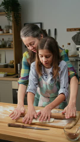 Medium vertical shot of teenage Caucasian boy in apron rolling out piece of dough on table for apple pie and chatting to mom, while baking pastry together at home at weekend