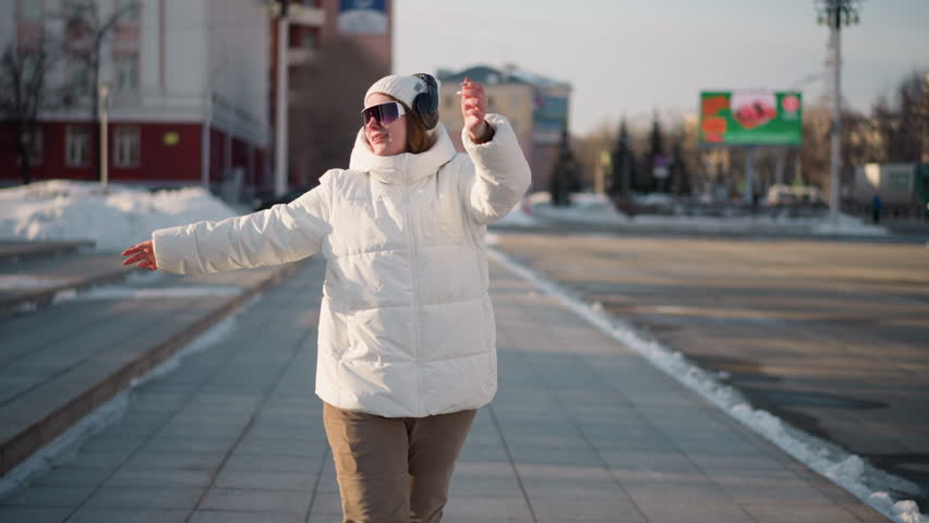 Young woman gliding along city sidewalk with arms raised, moving to rhythm in winter attire, wearing headset and sunglasses, joyful expression while crossing urban street with snow piles