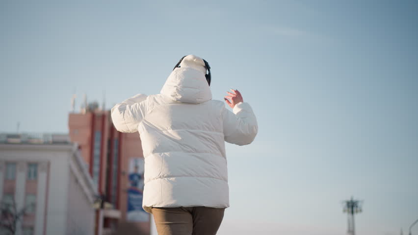 Cheerful dancer wearing white winter coat and beanie moves rhythmically in open urban space, back turned to camera, enjoying winter atmosphere under clear sky with city buildings in background