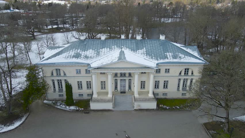 Gorgeous aerial drone footage view descending and flying lower in front of a large white and historic Saku manor mansion or Saku Mõis during a winter cloudy day, with leafless trees and snow around it