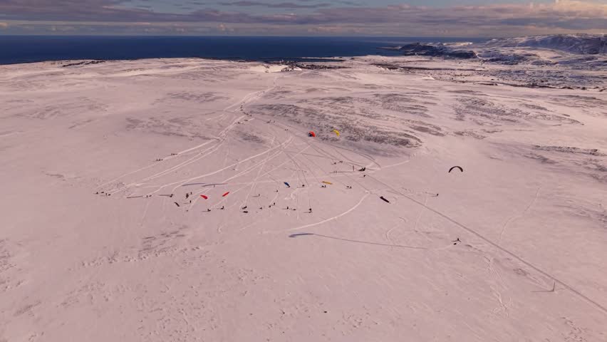 Gorgeous orbiting aerial drone footage spinning around people who are kite skiing or surfing on white snow covered Norway arctic landscapes while large mountains and Bering or Barents sea is in back.
