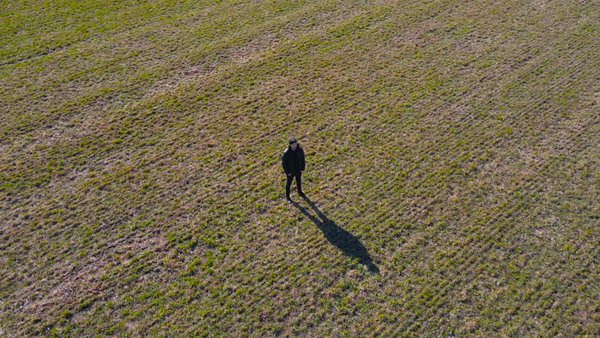 Aerial drone footage orbiting and spinning around a man with black clothing and a hat who is standing alone and still in a green and brown farmland or field during a sunny spring day, creating shadow.