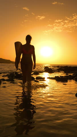 SILHOUETTE, LENS FLARE, VERTICAL: Woman in a bikini holds a surfboard as she walks toward camera through shimmering shallow water at sunset. Surfer is heading back to beach after sunset surf session.