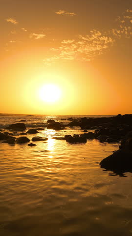 SILHOUETTE, LENS FLARE, VERTICAL: Rising sun and a surfer heading towards breaking waves in golden light. Woman in bikini and with surfboard in her hand walks between rocks in glistening shallow water