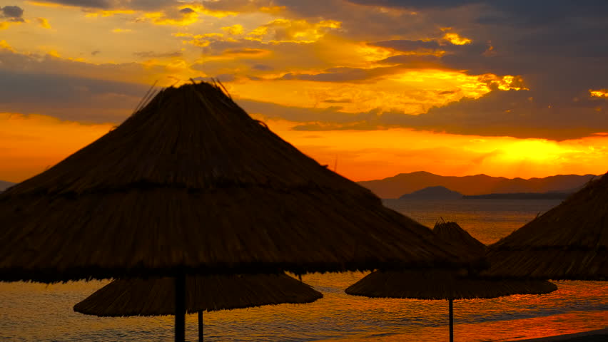 Sun setting behind straw umbrellas on beach. Golden hour sunlight bathes clouds in warm hues behind straw umbrellas on a tropical beach, creating a serene atmosphere perfect for relaxation