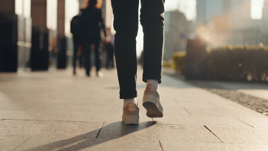 Close-up back view gimbal following shot female feet steps walking busy sidewalk, sunny day, young woman wearing black pants and white sneakers goes city street. Low angle people legs stepping avenue