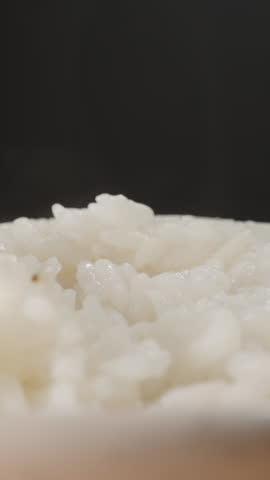 Vertical video. Wooden Spoon Lifting Steaming Freshly Cooked White Rice from a Bowl, Showing Soft Texture and Moisture Rising in Warm Steam Against a Dark Background