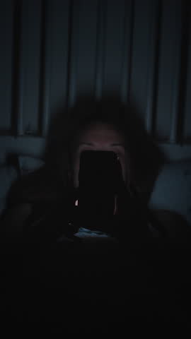 Vertical video. Woman Lying in Bed at Night Looking at Smartphone Screen in Complete Darkness with Only Screen Light on Her Face, Front Angle, Insomnia Concept