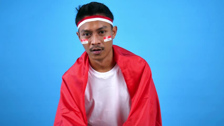Joyful young man with painted face and headband, wearing a white t-shirt, waving a red and white flag against a vibrant blue background. Celebration and patriotism concept.