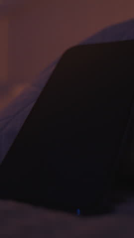 Vertical video. Woman Reaches to Pick Up Smartphone with Ringing 6 AM Alarm While Lying in Bed in Dim Light, Close-Up Low Angle, Sleepy Morning Concept
