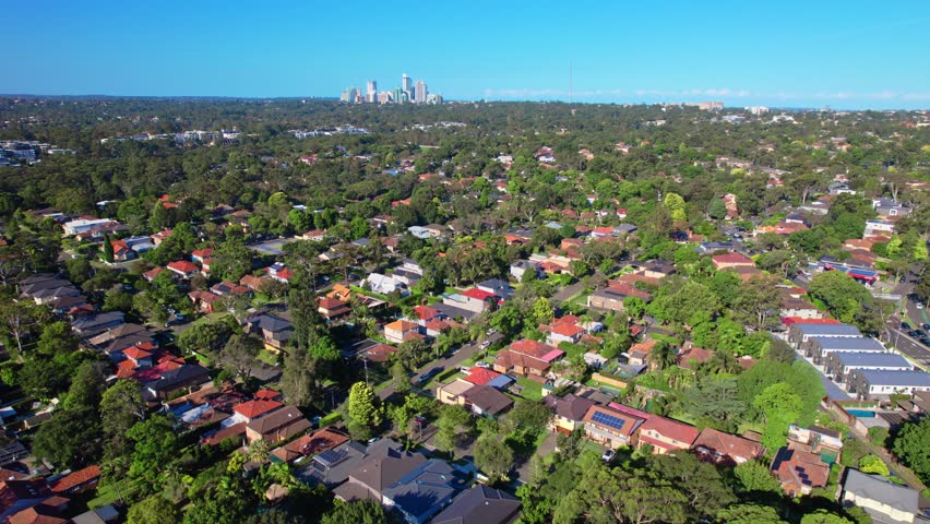 Aerial view of a peaceful suburb near a big city. Features single-story houses, tree-lined streets, and light traffic with distant skyscrapers visible. Bright summer day with lush greenery.