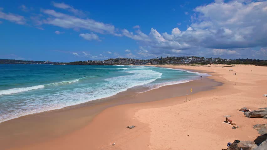 A stunning drone flight over Sydney's Curl Curl Beach on a bright summer day reveals mesmerizing ocean waves, turquoise waters, golden sand, and cloud-dotted blue skies.