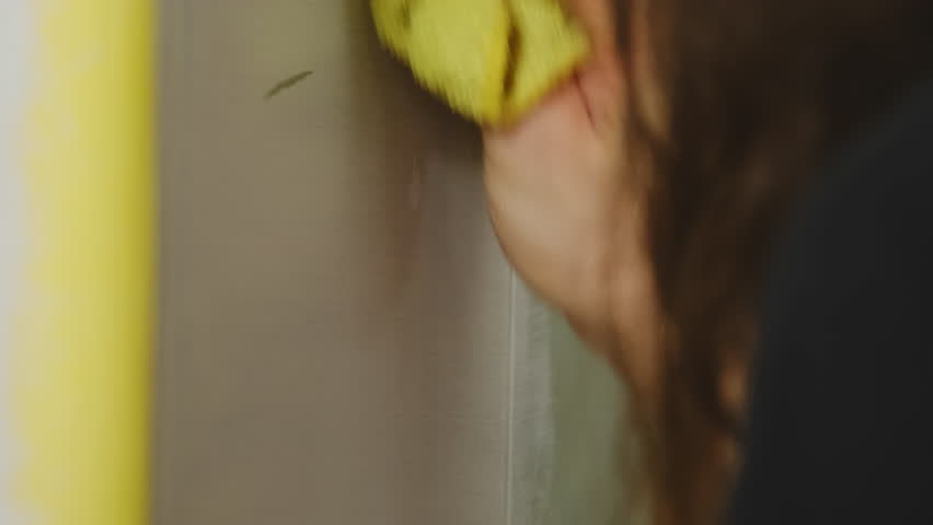 Woman wipes wall with damp rag removing smudges in room. Housewife carefully cleans surfaces to maintain neat and tidy living environment