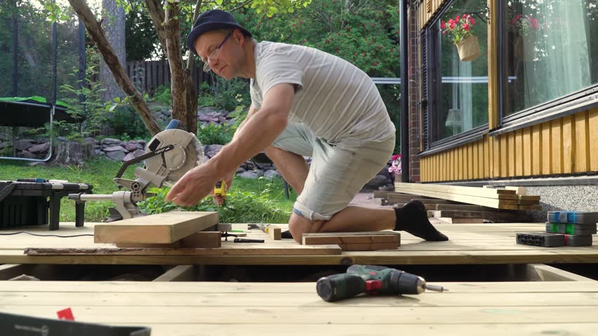 Man working on wooden deck construction outdoors using a circular saw, building a patio in backyard on a sunny day.