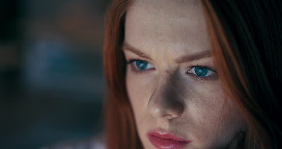 A closeup of a thoughtful young woman with vibrant red hair and striking blue eyes, showing deep focus.