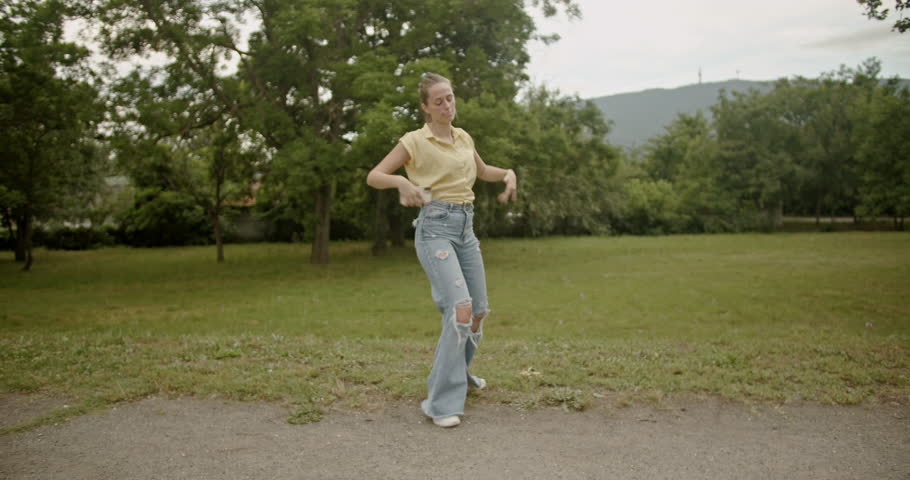 A young woman dances freestyle in a park, enjoying the music from her phone. She expresses freedom and happiness through her movements.