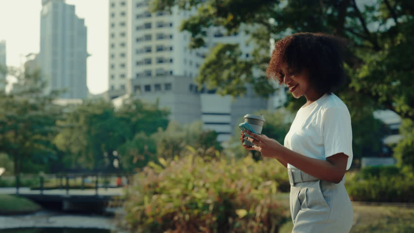 Positive African American woman carries smartphone and coffee cup walking across urban park. Young adult female reads chat on mobile phone outdoor office buildings side view