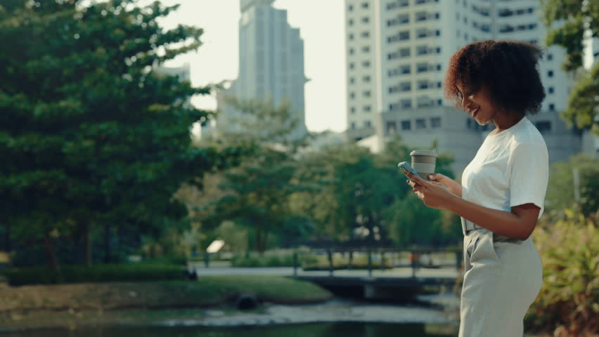 Positive African American woman carries smartphone and coffee cup walking across urban park. Young adult female reads chat on mobile phone outdoor office buildings side view