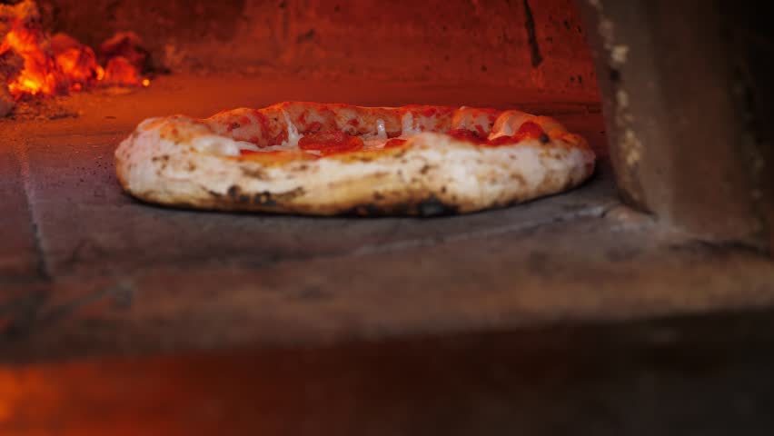 Traditional Italian pizza is cooked in a stone wood-burning oven. The chef takes the cooked pizza out of the oven with a long-handled spatula.