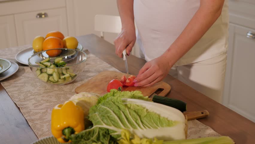 Close-up of a pregnant woman cutting a fresh tomato on a chopping board, she puts tomatoes in a plate. Preparation of vegetable salad.