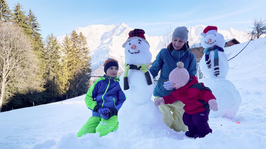 Family, mother two kids build together snowman dressed in hat scarf, outside in with snow mountain on background