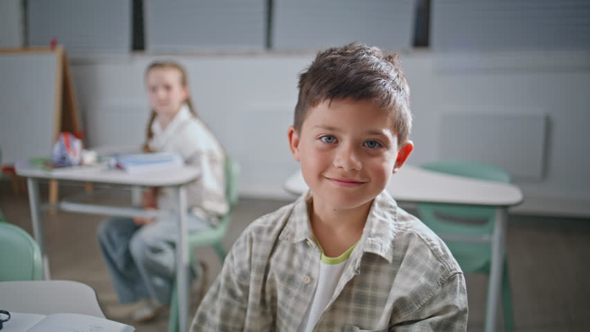 Portrait small kid sitting lesson in classroom. Smiling schoolboy listening teacher looking camera at cabinet closeup. Little happy boy learning in school auditorium. Joyful student schooling at desk
