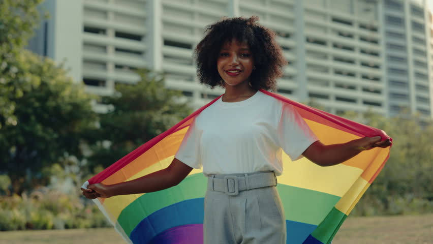 Portrait of smiling freedom woman wrapping with rainbow flag standing near commercial building. Positive African American female proud of LGBT community in urban area. Pride month