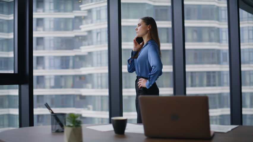 Confident woman dialing phone number at luxury company office. Serious businesswoman calling partners waiting answer at workspace. Elegant lady brunette standing at panoramic window looking city view.