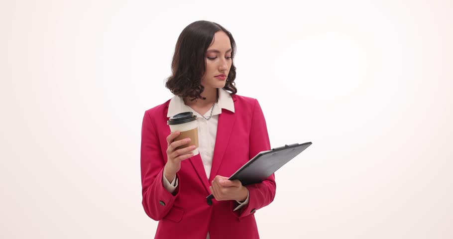 Woman in pink blazer drinks from disposable coffee cup holding black clipboard and reading document. Female professional takes break and multitasking
