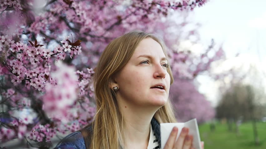 A woman experiences allergy symptoms and sneezes while standing under a beautiful cherry blossom tree in full bloom. The vibrant pink flowers represent the beauty of spring and the challenge of