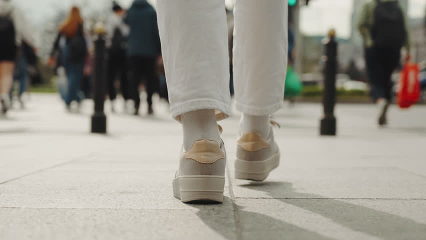 Back view following shot female feet steps walking pedestrian crossing street, sunny day, young woman wearing white pants and sneakers goes crosswalk. Low angle people legs stepping zebra asphalt