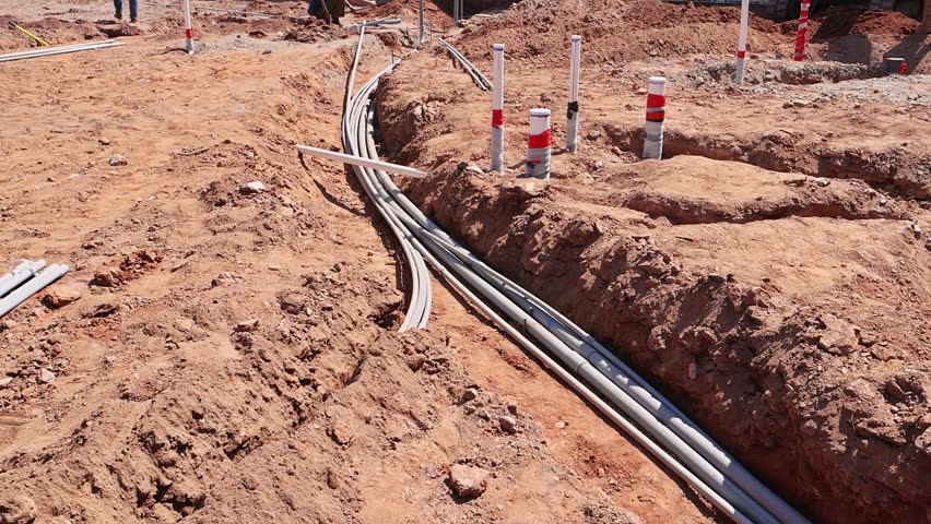 Workers organize electrical conduits in trench at construction site during foundation work