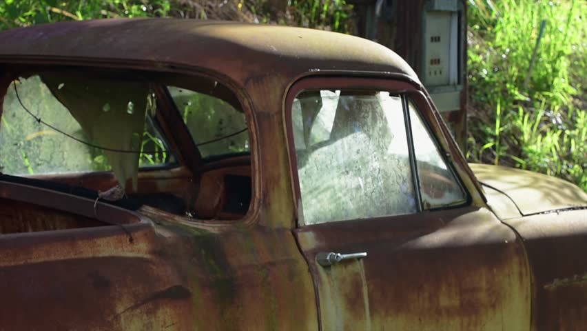 close up of and old abandoned wreck of a 1950s Holden FC utility pickup, rusted and lying in the undergrowth at the edge of a quiet lane in the morning sunshine, with an old petrol bowser