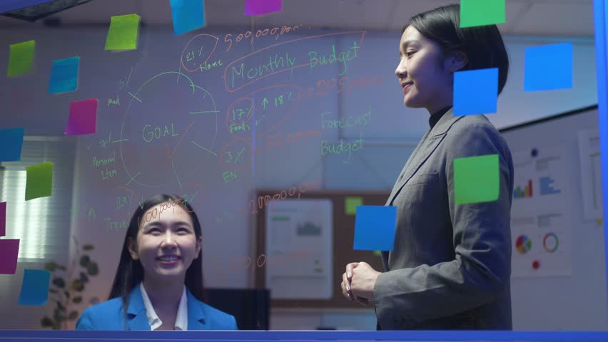 Two women are standing in front of a white board with colorful stickers on it. They are smiling and seem to be discussing something. The board has a lot of writing on it, including numbers and words