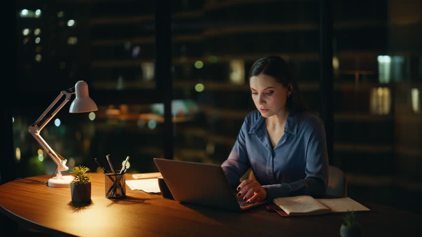 Hardworking freelancer working online at dark apartment workplace. Busy woman professional typing laptop reading notebook in night office. Overtime manager holding coffee cup thinking on deadline.