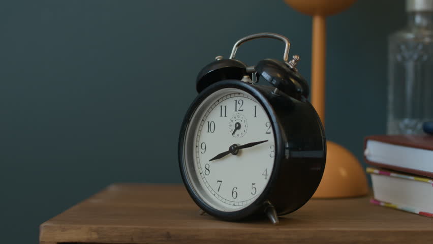 Close up view of hand of unrecognizable man reaching to turn off black vintage alarm clock placed on wooden nightstand in bedroom