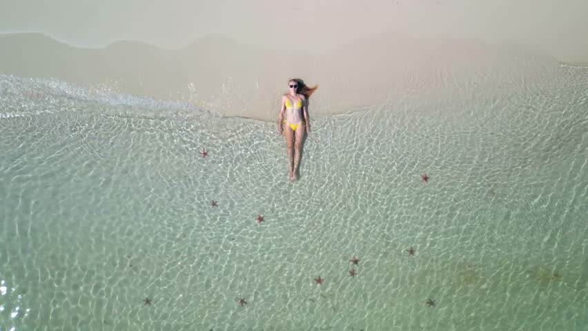 Aerial top-down view of woman in yellow swimsuit relaxing in clear turquoise sea with starfish on tropical white sandy beach on Phu Quoc Island, Vietnam. Concept of vacation, summer, and freedom.