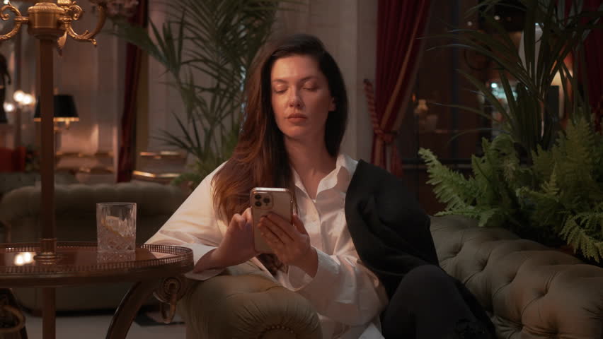 Businesswoman sitting on couch using smartphone in elegant lounge, wearing white shirt and blazer, relaxed posture, surrounded by plants and warm lighting, working or browsing during evening time