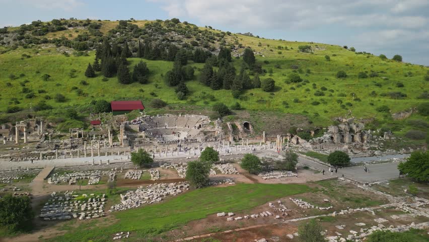 Aerial photo of Ephesus' small amphitheater in Turkey. Shows side perspective with adjacent ruins and green hillside. Historic columns line the foreground, telling ancient stories.