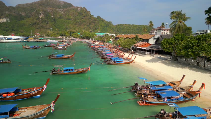 Aerial view of colorful traditional longtail boats anchored along white sand beach with turquoise water on Phi Phi Island, Thailand.