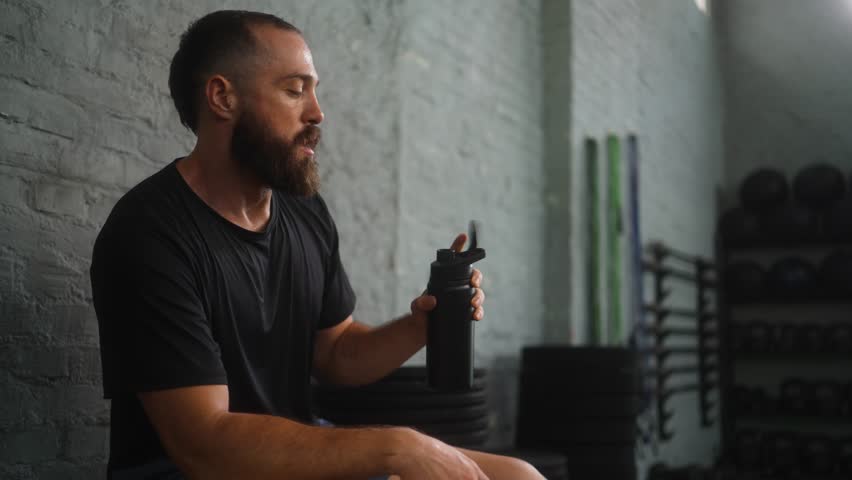 Athletic mid adult bearded man drinking water from black tumbler bottle after intense workout in modern fitness facility with white brick walls demonstrating proper hydration