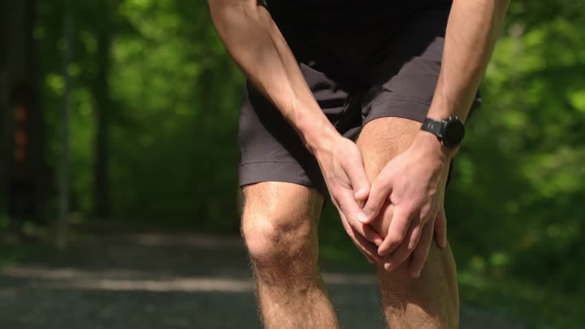 Runner holding knee in pain during workout in summer park. Man has injury while running. Male athlete suffering from knee pain after sport exercises